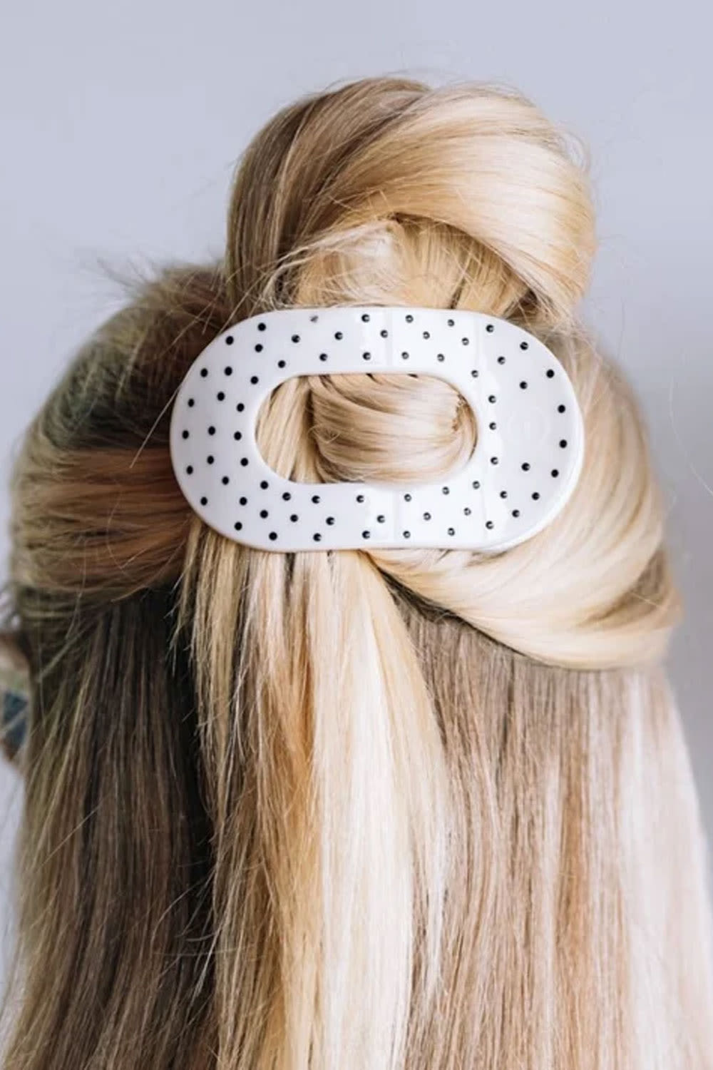 A white oval hair claw clip with black polka dots, shown holding a blonde and brown ponytail hairstyle.