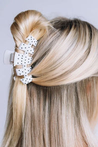 A close-up of a blonde person's hair styled with a white claw clip featuring black polka dots, shown from the back against a light gray background.