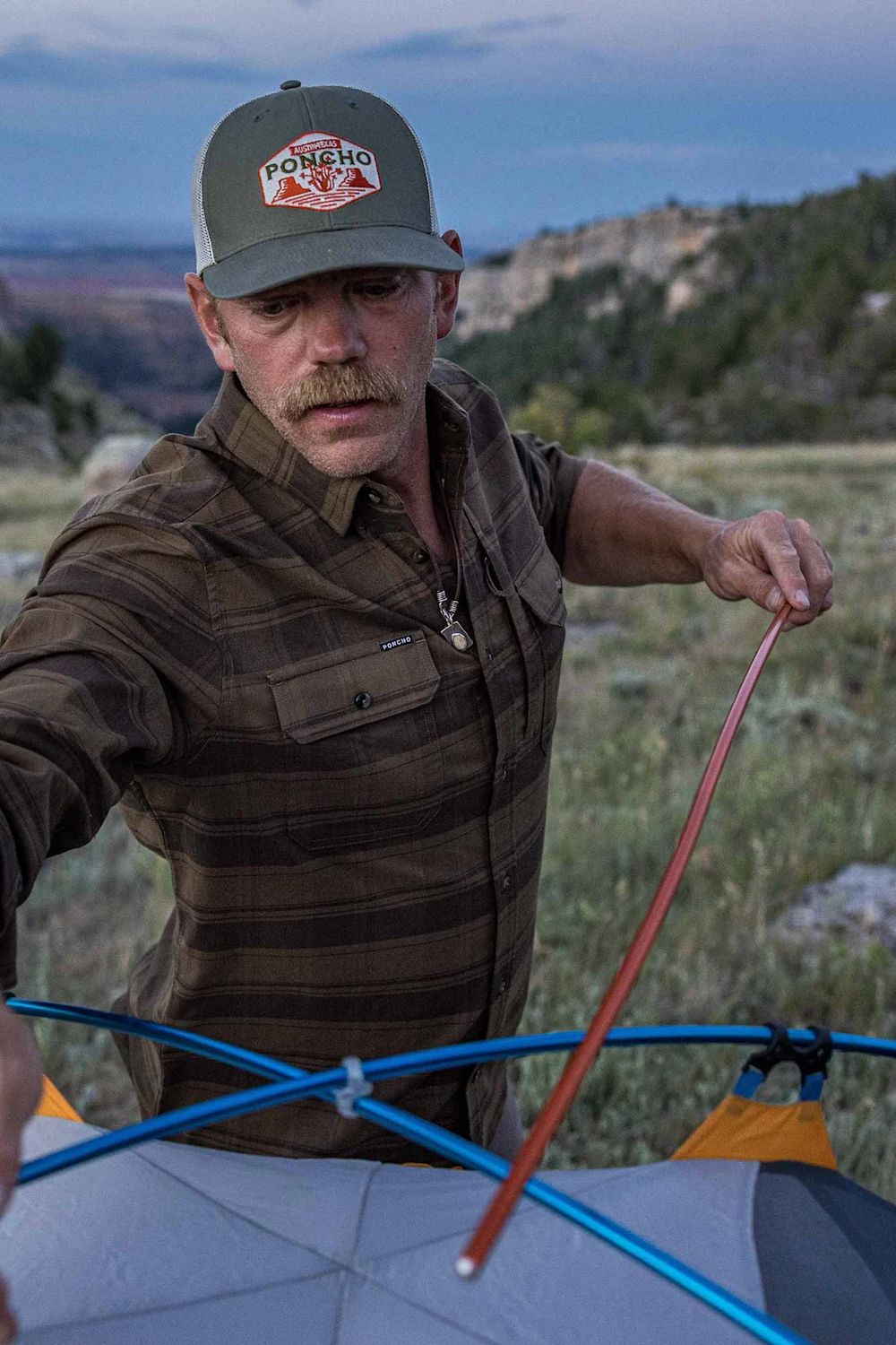 A man outdoors in a mountain landscape wearing a brown plaid shirt and a gray Pongho cap, holding tent poles above a gray tent.