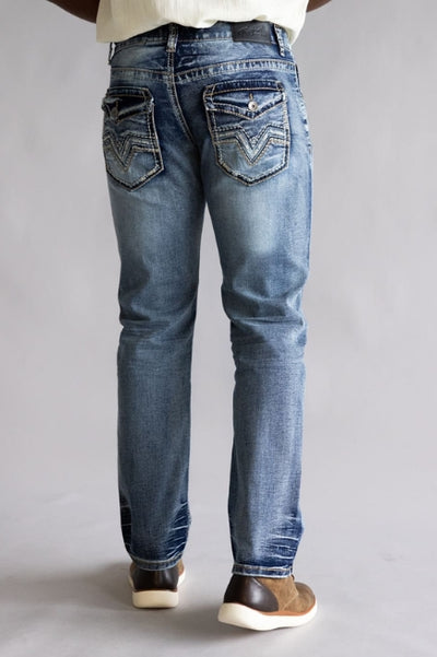 Back view of a model wearing medium-wash straight-leg jeans with decorative stitched flap back pockets, paired with a white top and brown slip-on shoes against a neutral gray background.