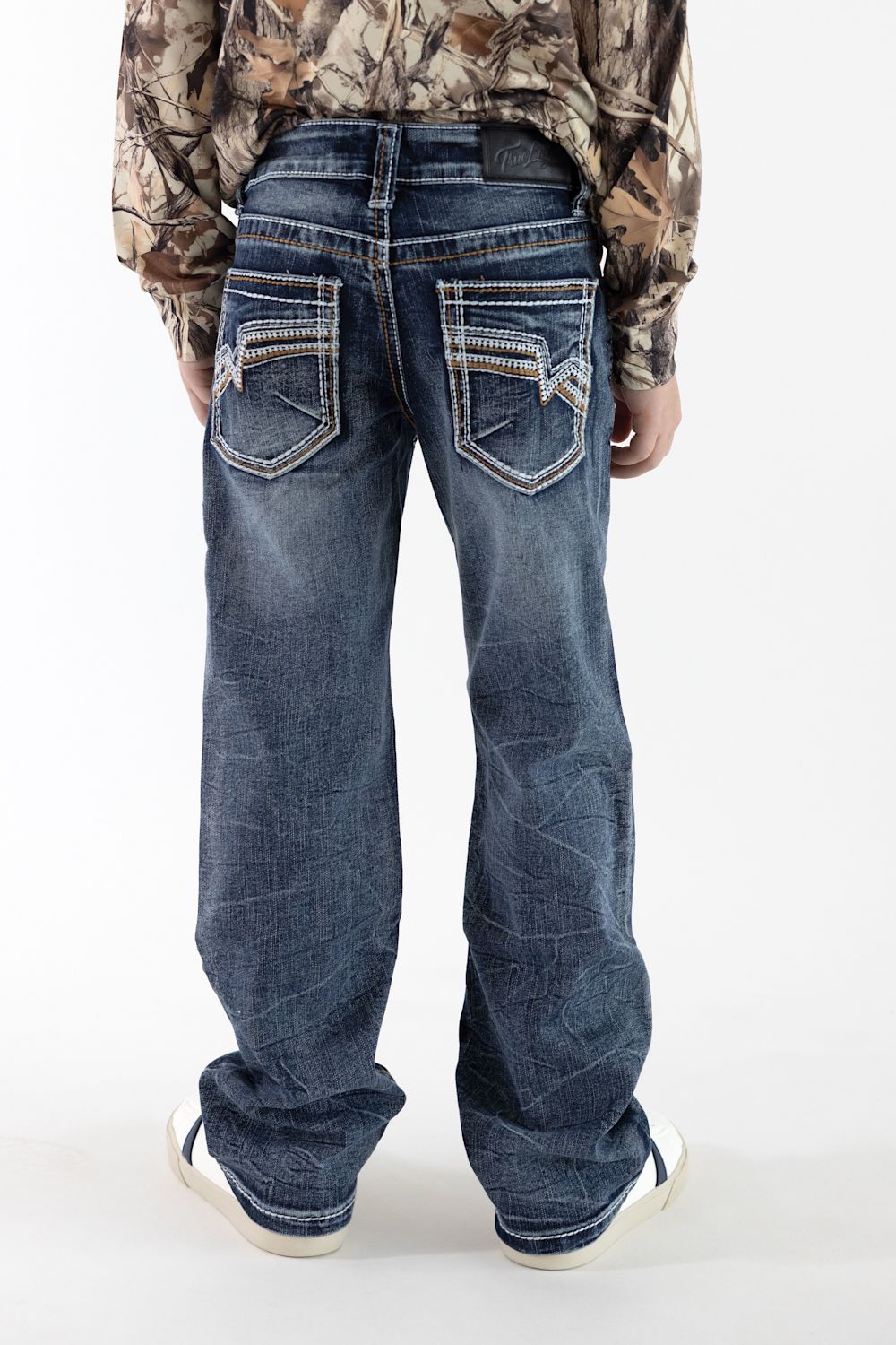A boy viewed from the back wearing dark wash denim jean with detailed stitching on the back pockets, a long sleeve camo shirt, and white sneakers in front of a white studio background