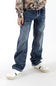 A boy wearing dark wash denim jeans with a long sleeve camo shirt and white sneakers in front of a white studio background