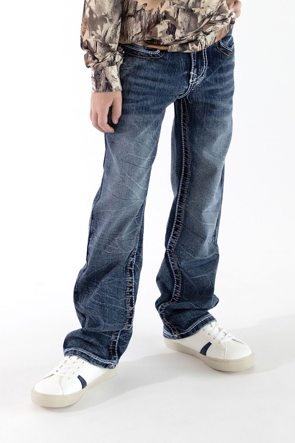 A boy wearing dark wash denim jeans with a long sleeve camo shirt and white sneakers in front of a white studio background