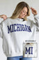 A woman wearing a light heather gray sweatshirt with 'MICHIGAN' printed in navy blue across the chest. The image includes a small inset showing the reverse side of the sweatshirt with 'MI' printed in navy.