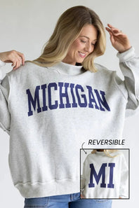 A woman wearing a light heather gray sweatshirt with 'MICHIGAN' printed in navy blue across the chest. The image includes a small inset showing the reverse side of the sweatshirt with 'MI' printed in navy.