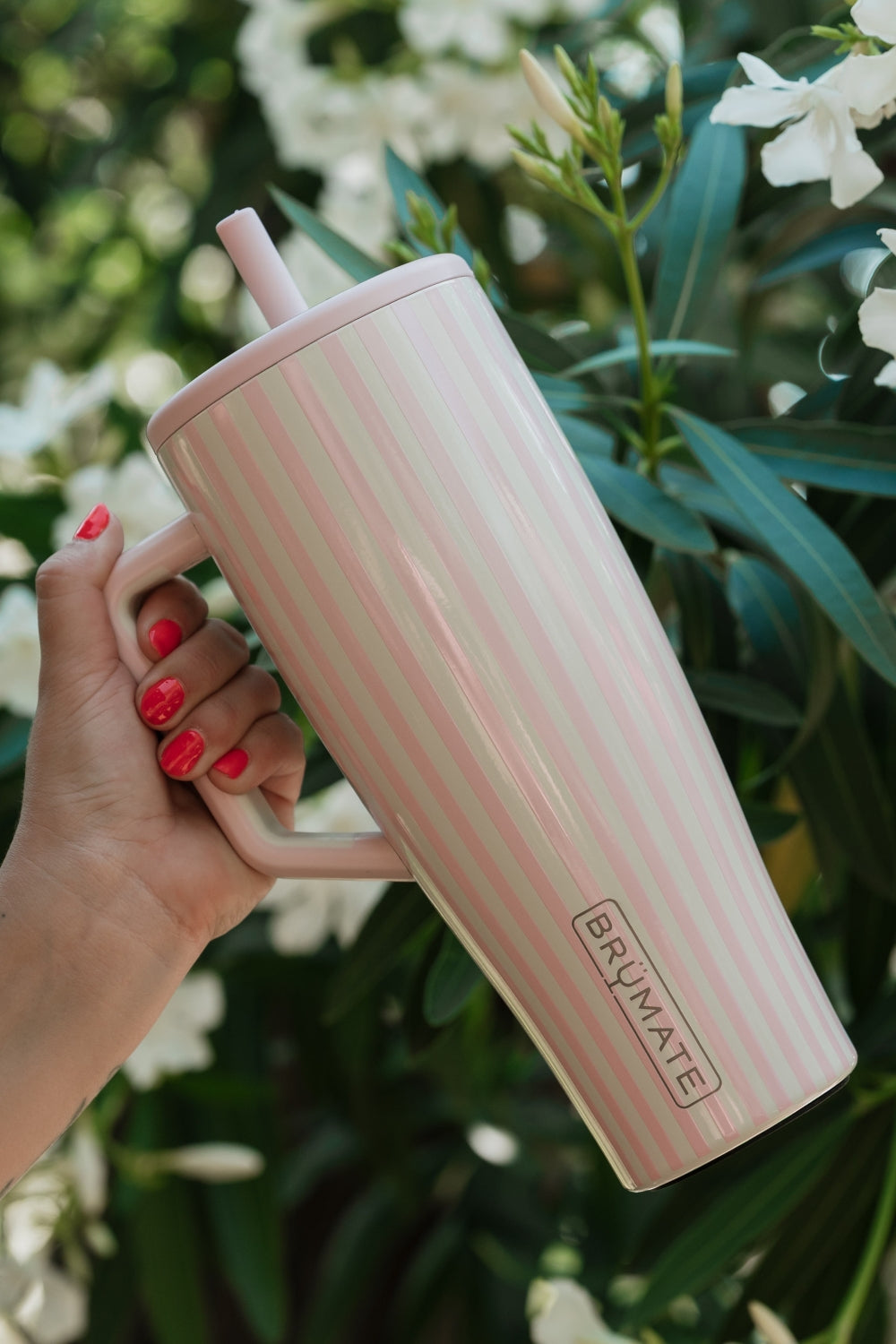 A hand with red nail polish holds a tall, tapered travel tumbler with a handle and straw. The tumbler has a light pink and white vertical stripe pattern and features the brand name 'BRuMATE' printed near the bottom.