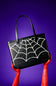 A black tote bag with a bright green glow in the dark spiderweb pattern printed across the front, featuring two black shoulder straps, set against a plain white background.