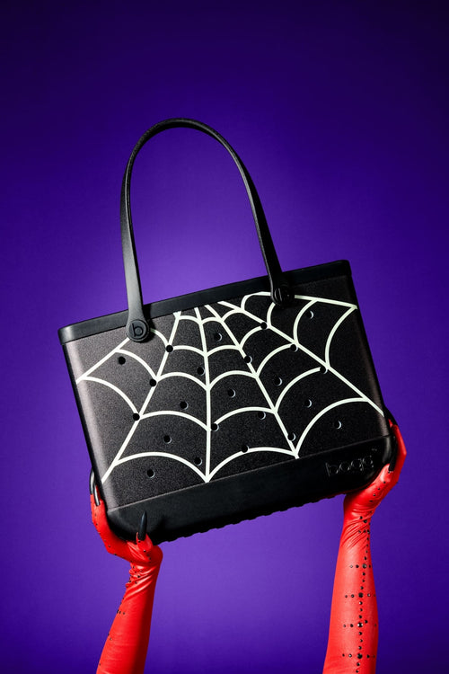 A black tote bag with a bright green glow in the dark spiderweb pattern printed across the front, featuring two black shoulder straps, set against a plain white background.