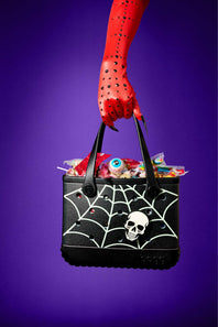 A black Halloween-themed tote bag with a white spiderweb pattern and skull graphic, held by a red arm against a purple background. The bag is filled with candy and is decorated with small red rhinestones.