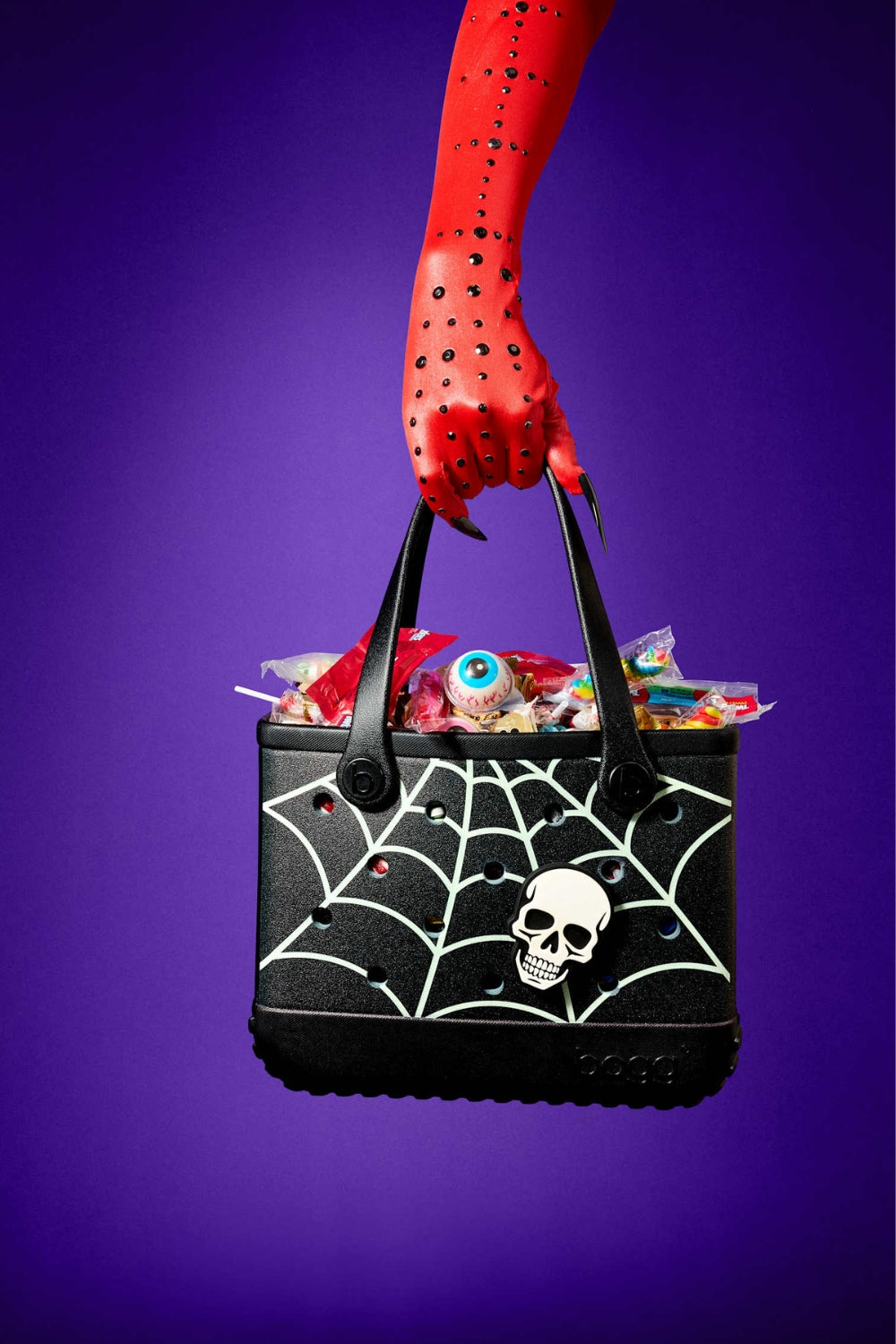 A black Halloween-themed tote bag with a white spiderweb pattern and skull graphic, held by a red arm against a purple background. The bag is filled with candy and is decorated with small red rhinestones.