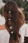 A woman viewed from behind with long, wavy brown hair styled in a half-up half-down braid secured with four decorative hair clips in different metallic and textured designs. She is wearing a white off-the-shoulder top, with a soft-focus outdoor background of trees and light-colored rocks.
