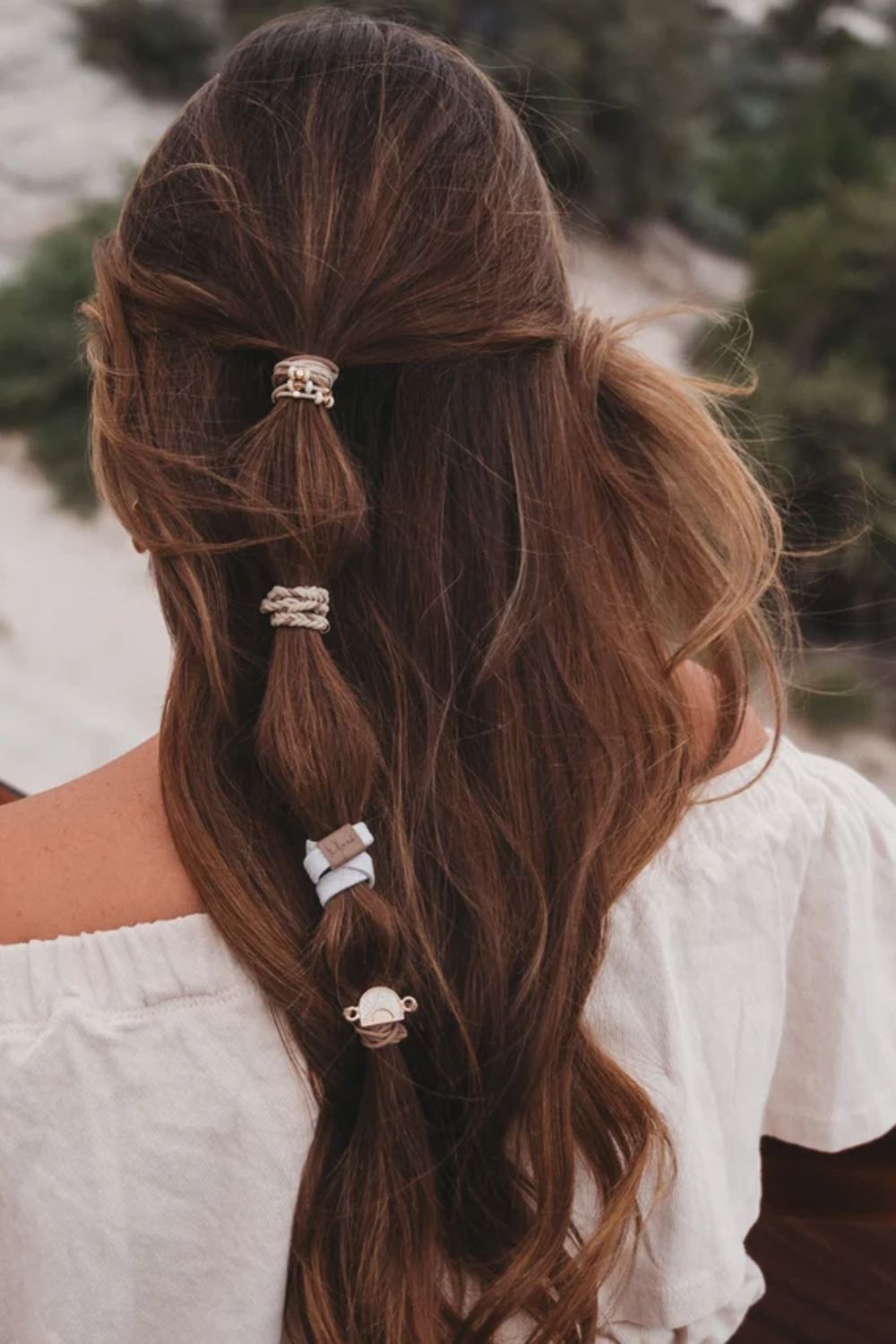 A woman viewed from behind with long, wavy brown hair styled in a half-up half-down braid secured with four decorative hair clips in different metallic and textured designs. She is wearing a white off-the-shoulder top, with a soft-focus outdoor background of trees and light-colored rocks.