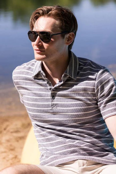 Man wearing a blue/grey and white striped polo shirt and black sunglasses sitting on the sand by a lake