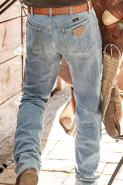 Person viewed from the back standing on a brick sidewalk wearing medium wash jeans and brown belt carrying a horse saddle