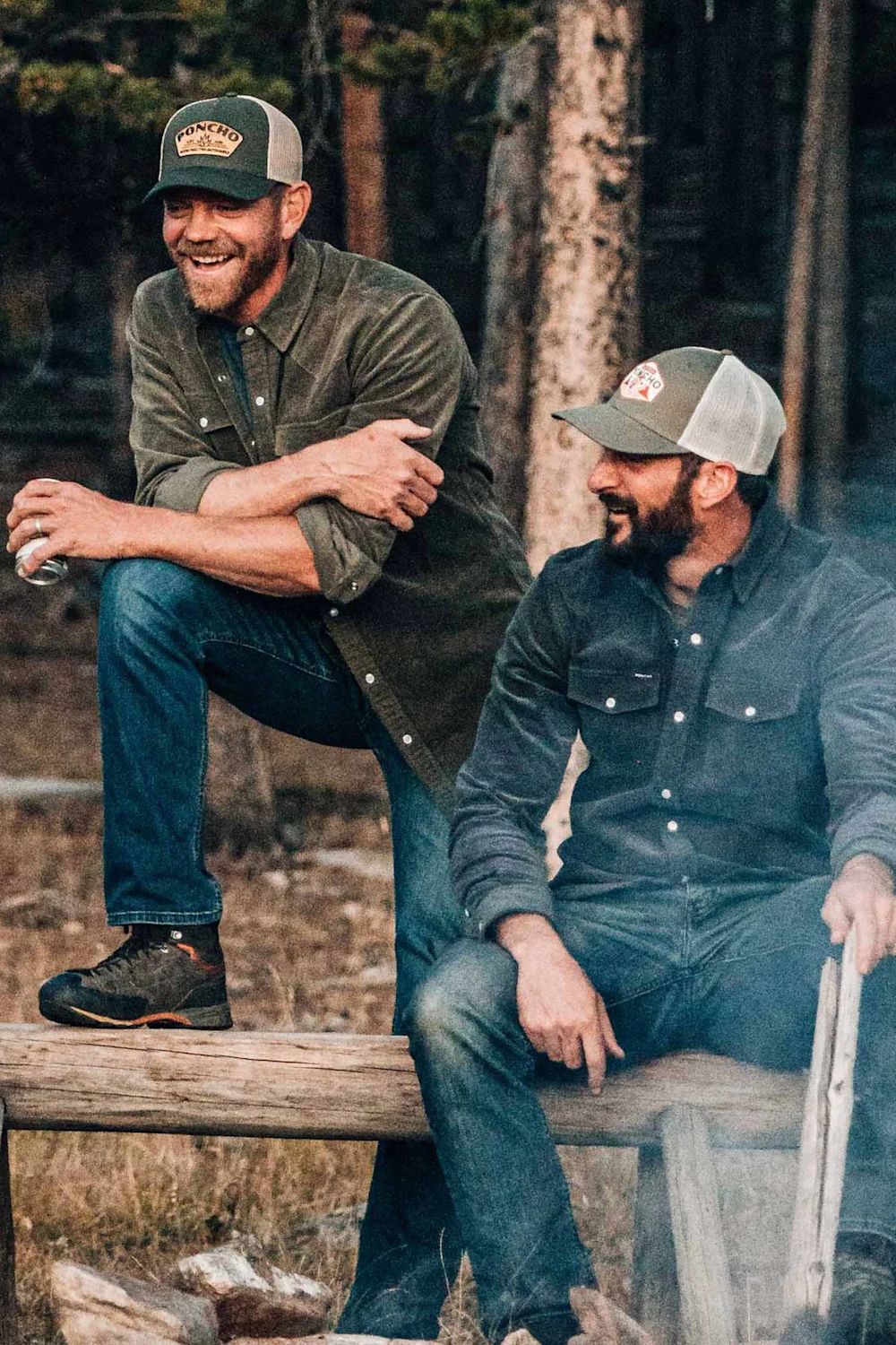 Two men in outdoor apparel sitting on a wooden bench in a forest setting. One man wears a green button-up shirt and blue jeans, while the other wears a dark gray button-up shirt and blue jeans.